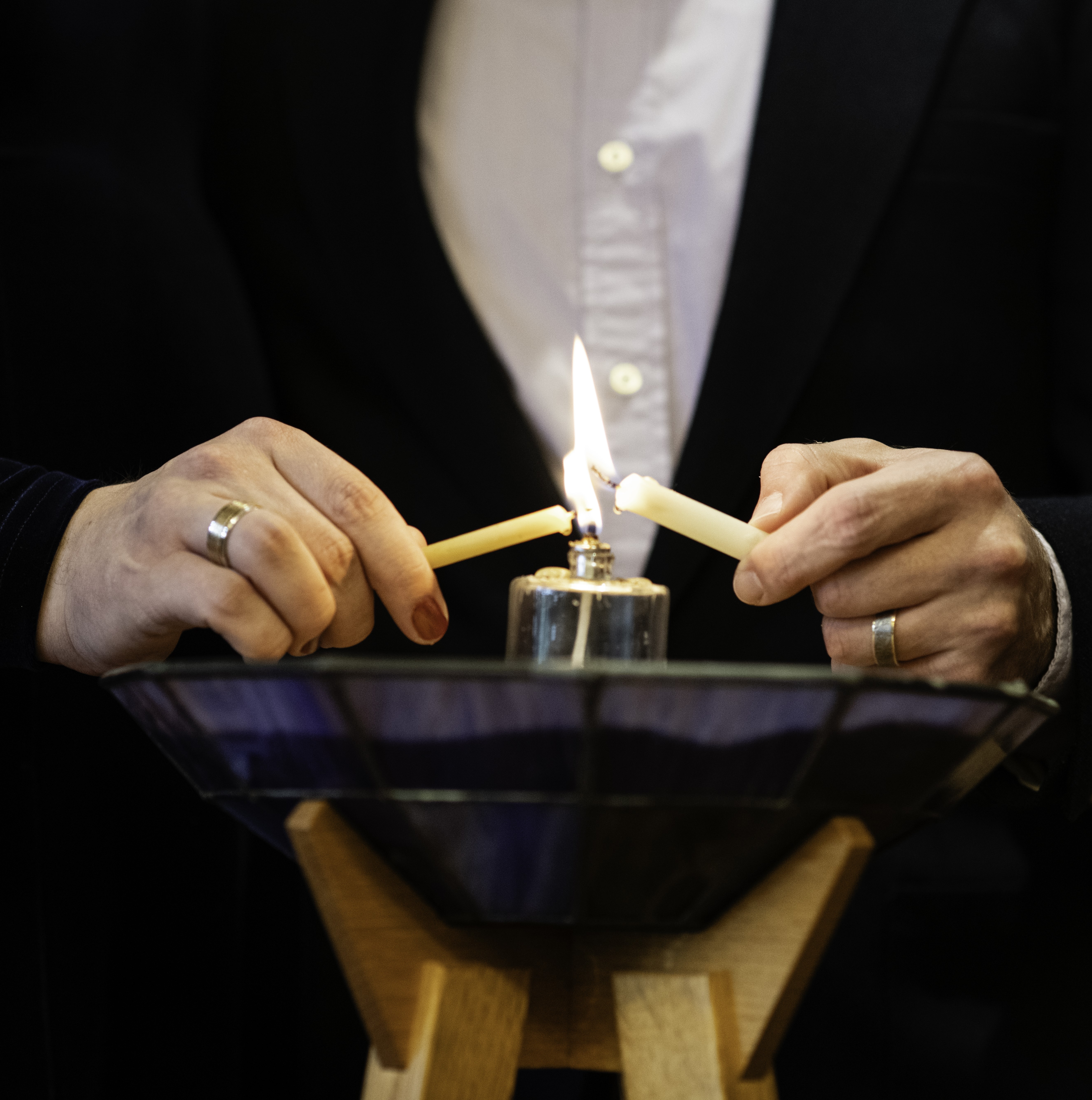 hands of two people (the site-author & their spouse) lighting a chalice; the left hand has red nail-polish. photo by Brittany Brooke Crow, 2023