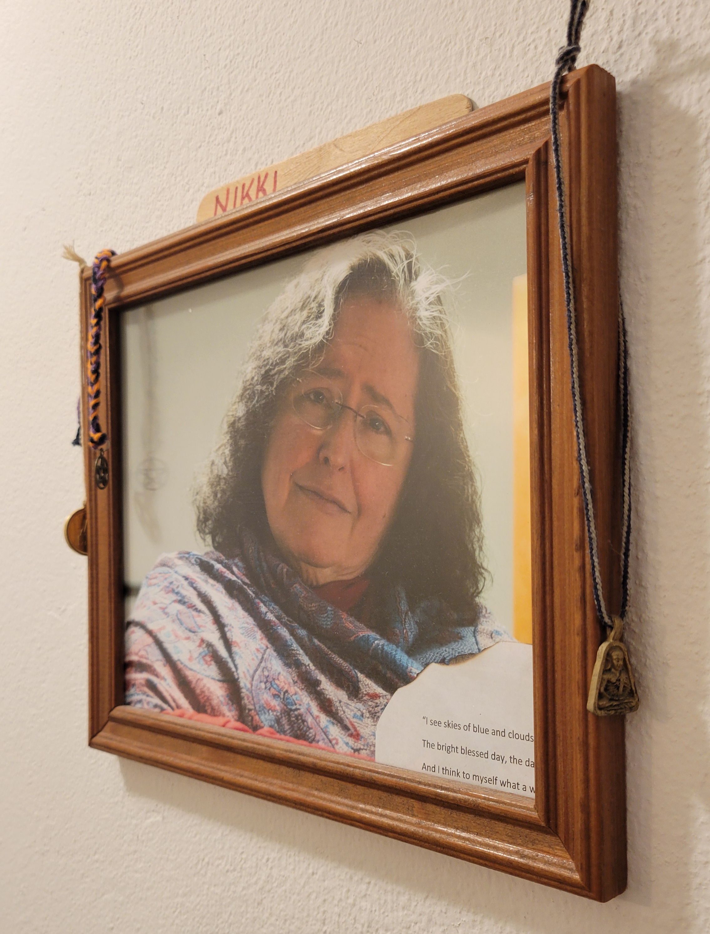 a framed photo of Nikki Bado hanging on the site-author's wall. there are pendants of the Buddha, slip of balsa wood with Nikki's name on it, and a braided cord with a pentacle