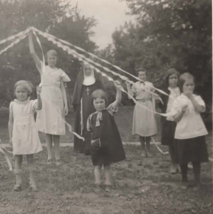 a found photo from the 1930s-1940s. 5 elementary-aged girls each hold ribbons of a maypole. a teenage girl stands in the center of the circle holding these ribbons up. a nun stands behind them all